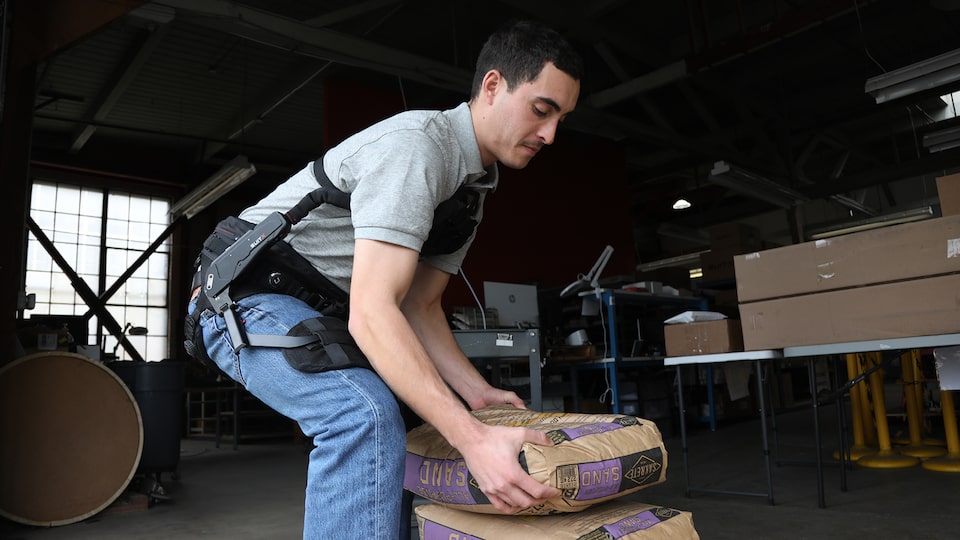 A man wearing a high-tech exoskeleton suit on his upper body bends down to lift a box in a warehouse.