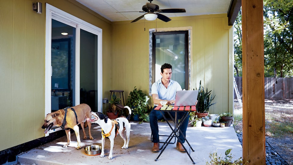 Architect Jon Hagar uses a latop while sitting at a table on a covered outdoor patio with his two dogs standing nearby.