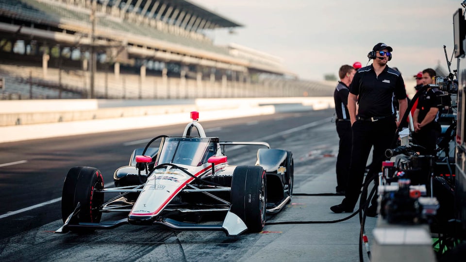 A race car is stopped on the track with crew and equipment nearby. The stands are visible in the background.