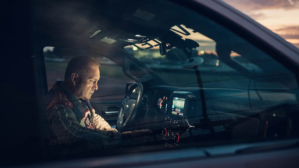 Tim Baughman sits in the cabin of a Chevrolet Silverado with the lights on as the sky is darkening outside.