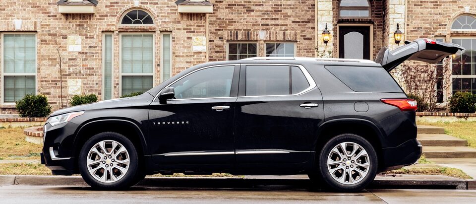 A black Chevy Traverse is parked curbside in front of a brick building with its back hatch open.