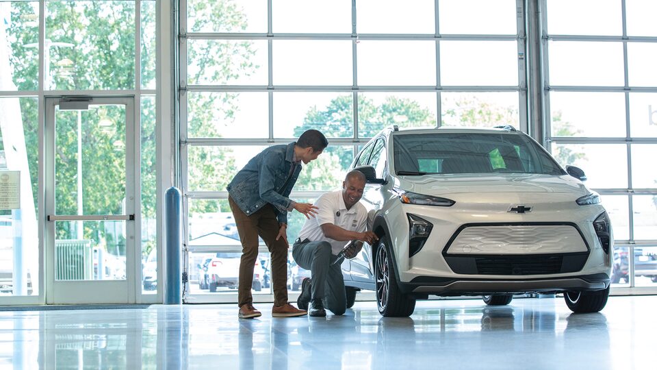 Dos personas conversan arrodilladas junto al neumático delantero de un Chevy Bolt en el taller de un concesionario de la marca.