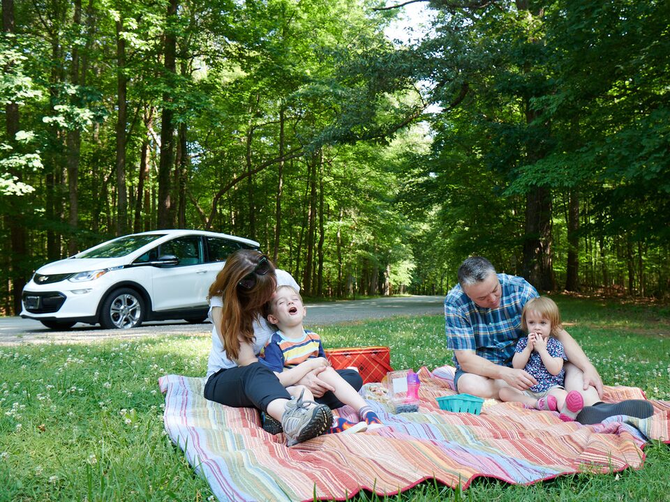 Un hombre y una mujer y dos niños están sentados sobre una manta colorida en el césped disfrutando un picnic, con un Bolt EV en blanco en el fondo.
