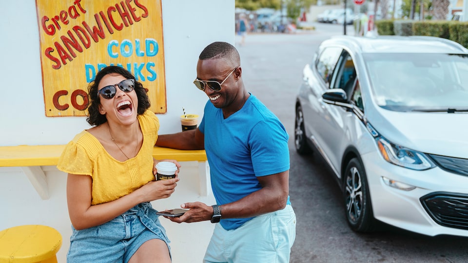 Un hombre y una mujer ríen juntos mientras se sientan en un mostrador frente a un cartel que dice "Great sandwiches, cold drinks". Hacia un lado se ve un Bolt EV en Plata Hielo Metálico estacionado.
