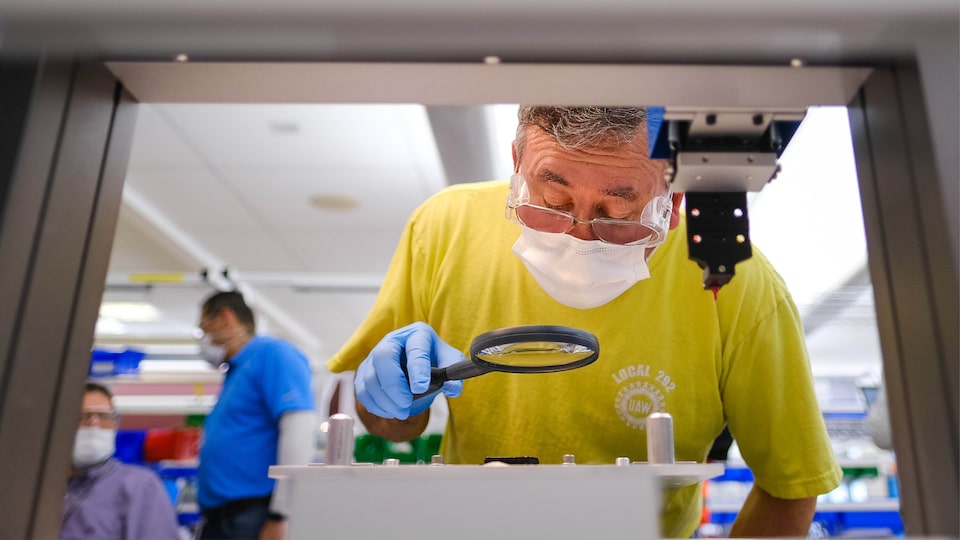 Un hombre con anteojos, mascarilla y guantes se inclina para examinar un equipo a través de una lupa.