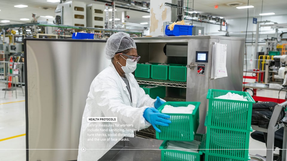 A woman in full personal protective equipment stacks bins of face masks outside a sanitizer, which is filled with more bins. Text on image reads: “Health protocols: Safety measures for workers include hand sanitizing, temperature checks, social distancing, and the use of face masks.”