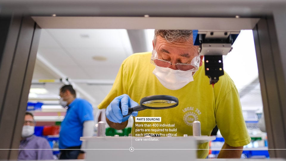 A man in a face mask leans down over a piece of equipment with a magnifying glass. Text on image reads “Parts sourcing: More than 400 individual parts are required to build each VOCSN V+Pro critical care ventilator.”