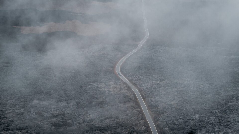 A drone shot of a strip of pavement winding through a field of black lava rock, seen through fog.