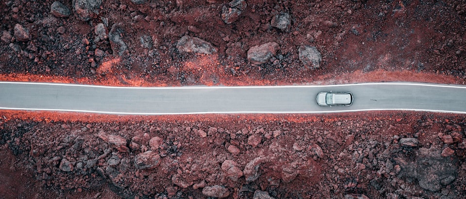 An overhead drone shot of a gray Chevrolet Blazer driving on a blacktop road through a field of lava rocks.