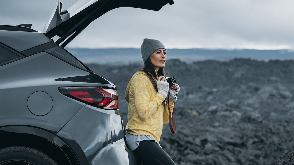 A woman wearing a sweater, hat and gloves holds a camera as she sits in the open tailgate area of a gray Chevrolet Blazer looking out at a surrounding field of black lava rocks.