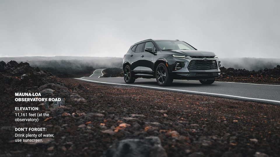 A gray Chevrolet Blazer drives on a narrow strip of pavement through a field of black lava rocks.