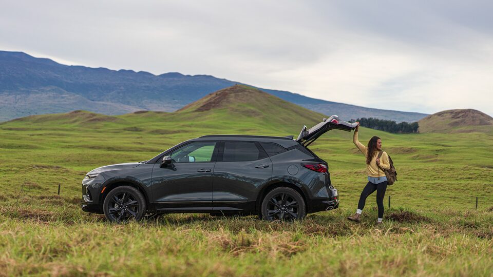 A woman stands behind a gray Chevrolet Blazer in an open, hilly field with mountains in the background. She has one hand on the open tailgate.