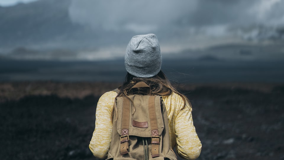 Seen from behind, a woman wearing a backpack and a hat looks out on a Hawaiian landscape of lava rocks, mountains, and clouds.