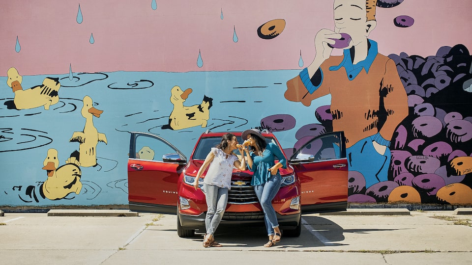 Two women lean against the front of a red Chevrolet Equinox while eating donuts. There is a mural on the wall behind them of a man eating a donut and ducks swimming in water.