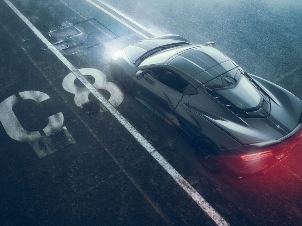 A miniature black Corvette Stingray seen from above, with a red glow coming from its taillights, appears to be moving on a backdrop of black pavement with lane markings and a white “C8” stencil next to the car.