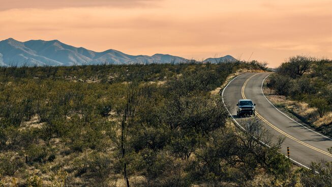 A black Tahoe RST drives down a winding road with mountains in the background. The setting sun has turned the clouds and sky behind the mountains a peach color.