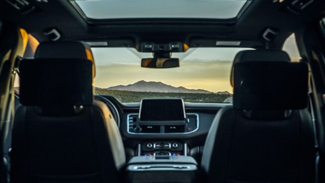 Seen from the back seat of a Tahoe RST, two people sit in the driver and front passenger seats at dusk. A mountain landscape spreads across the view out the windshield, and the sky is overhead through the open sunroof.