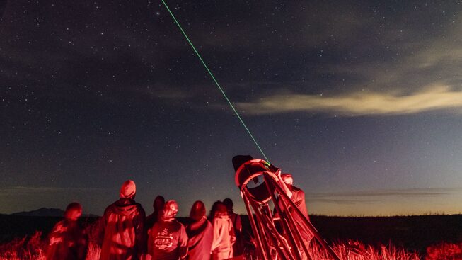 A group of people gather around a telescope that has a laser guide pointing into the darkening sky. The group are lit by red light to not interfere with night vision.
