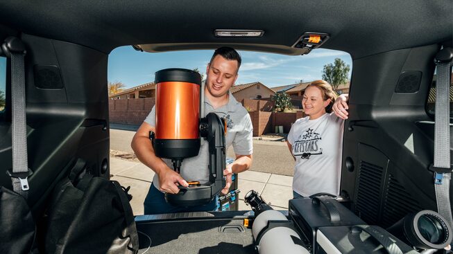 Seen from the interior of a Tahoe RST, Carlos Aragon and Kira Reid stand behind the open back hatch, loading telescopes into the cargo area.
