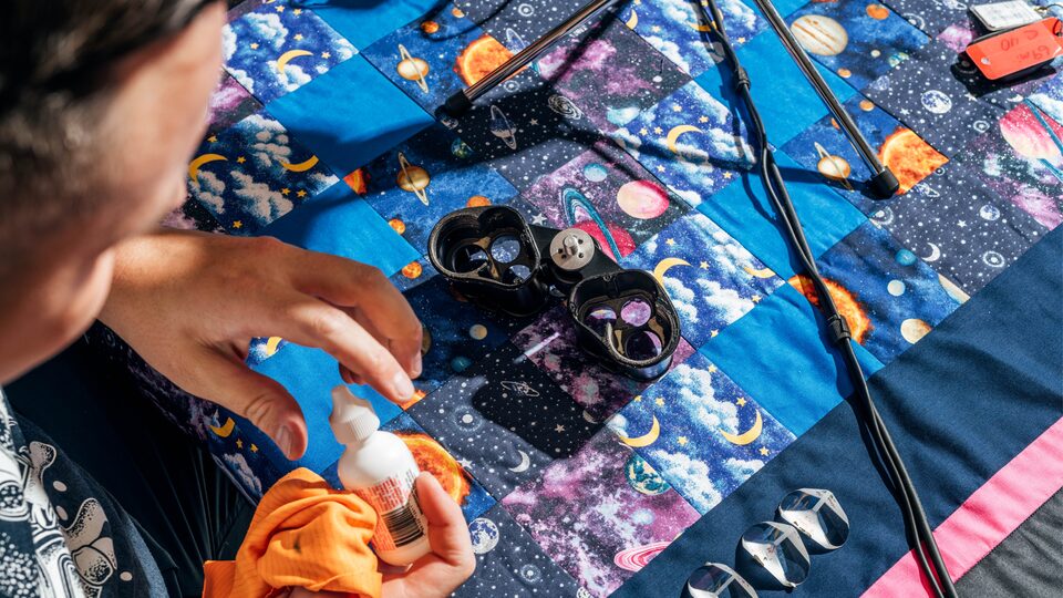 Carlos Aragon quita la tapa de una botella para preparar el mantenimiento de las piezas del telescopio, que se encuentran sobre una manta con temática astronómica frente a él.