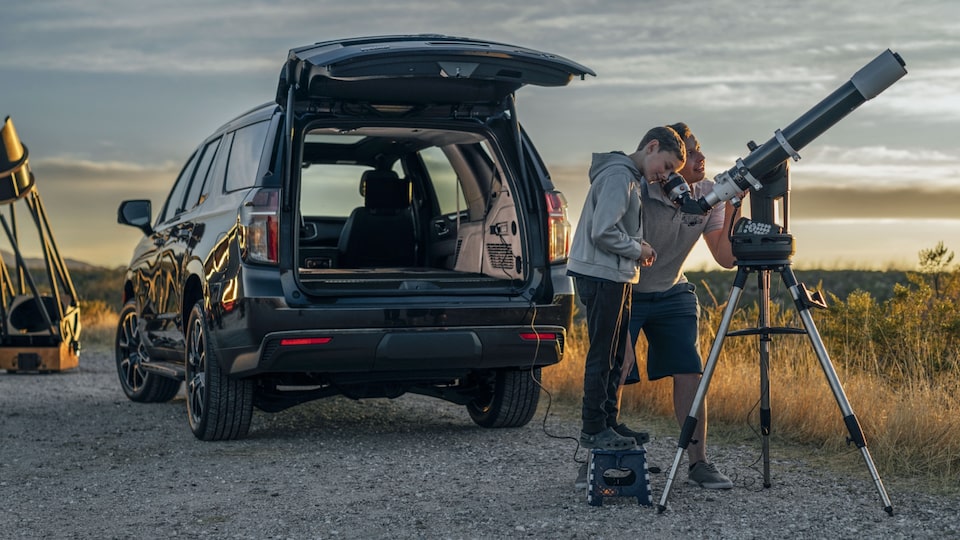 Carlos Aragon ayuda a un niño a mirar a través de un gran telescopio montado en un trípode detrás de una Chevy Tahoe en Negro mientras cae el atardecer sobre el paisaje desértico que los rodea.