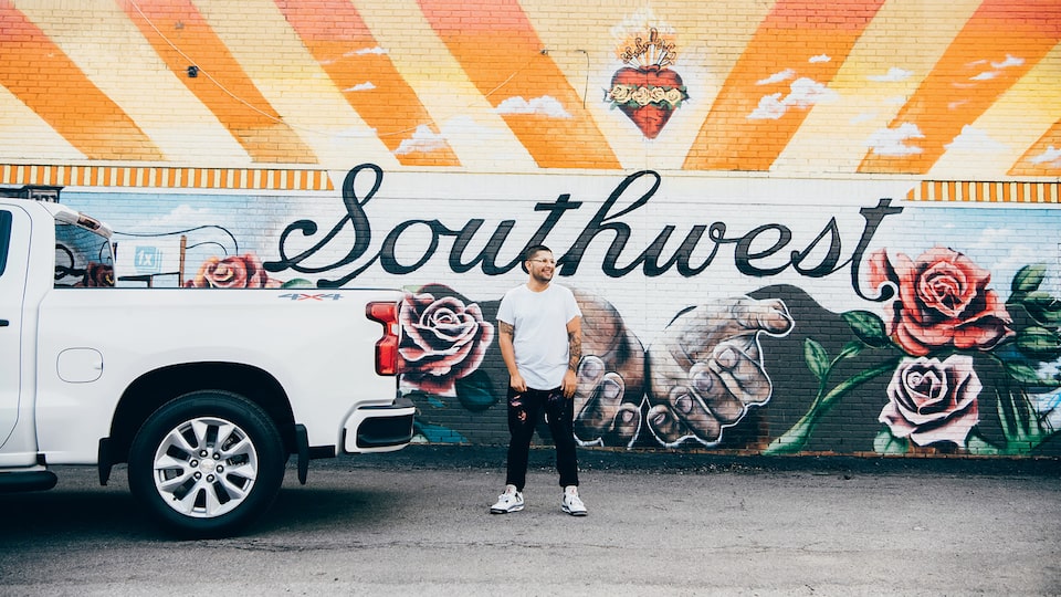 Freddy Diaz stands behind a white Silverado, in front of one of his colorful murals that features the word “Southwest,” a pair of hands, and roses.