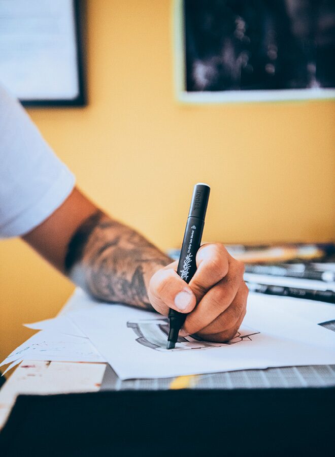 Freddy’s tattooed arm and hand are visible as he draws on some paper on a drafting board.