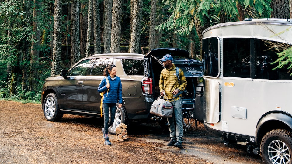 A young couple holding camping gear stand next to a 2021 Chevy Tahoe with an Airstream travel trailer hitched to it, parked among tall trees.