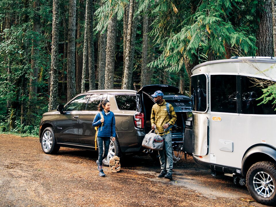 A young couple holding camping gear stand next to a 2021 Chevy Tahoe with an Airstream travel trailer hitched to it, parked among tall trees.