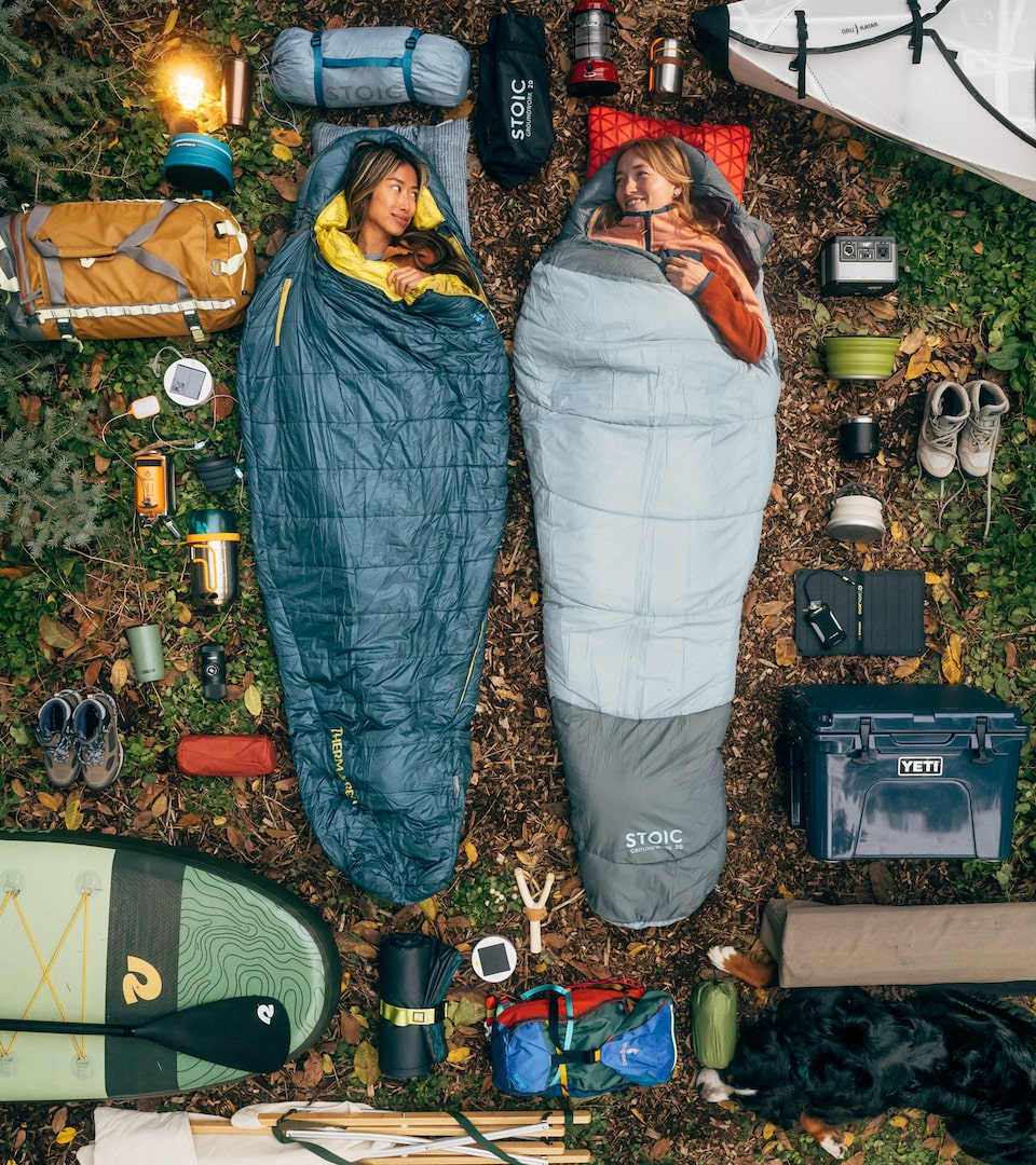 Vistas desde arriba, dos mujeres están acostadas en el suelo en un campamento, en bolsas de dormir rodeadas por una gran variedad de artículos de campamento.