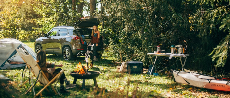 A Zeus Bronze Metallic Trailblazer ACTIV sits in a campsite surrounded by tall trees. Two people are in the campsite, along with various camping gear. A campfire burns in a small firepit in the center.