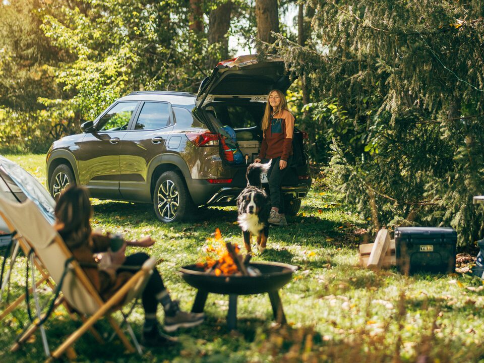 A Zeus Bronze Metallic Trailblazer ACTIV sits in a campsite surrounded by tall trees. Two people are in the campsite, along with various camping gear. A campfire burns in a small firepit in the center.