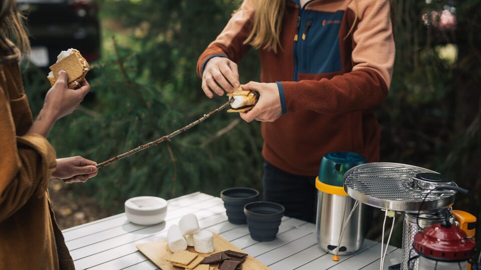 Two people stand at a table in a campsite making s’mores.