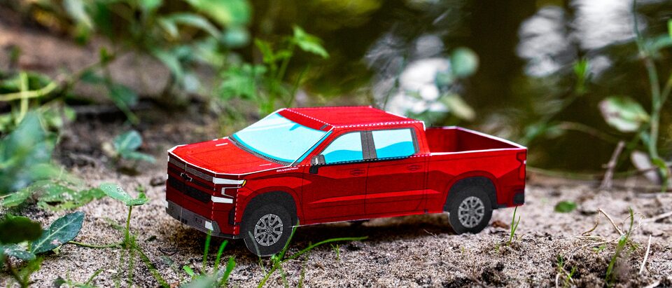 A miniature paper model of a Cajun Red 2020 Silverado RST Diesel sitting on bare ground with grass and leaves in the background.