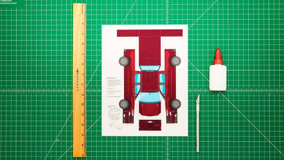 A paper template for the model Silverado sits on a green artist mat with an artist knife, a bottle of glue and a ruler. In the video, the paper truck is cut out and assembled and drives off the mat.