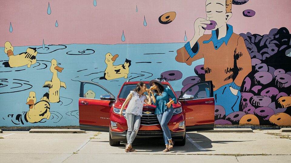 Two women lean against the front of a red Chevrolet Equinox while eating donuts. There is a mural on the wall behind them of a man eating a donut and ducks swimming in water.