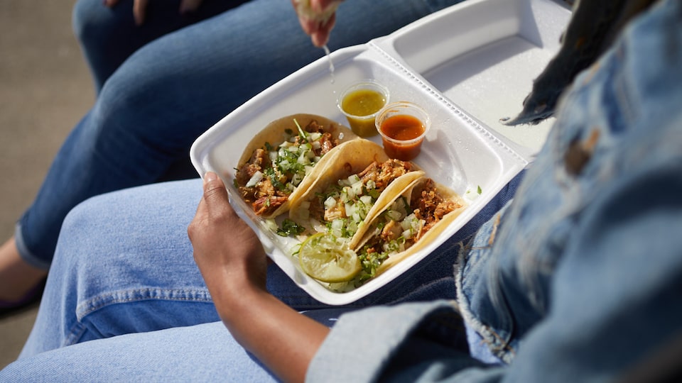 Three tacos and sauces sit in a large foam takeout box, which is on the lap of a person wearing jeans and a jean jacket.