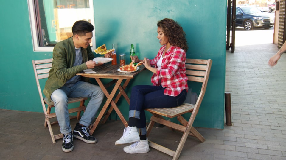 A young man and woman sit on folding chairs at a small table in an open-air restaurant eating pizza.