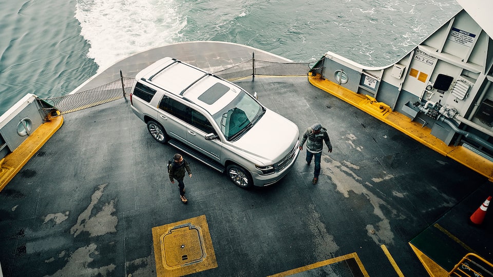 A father and son walking past a Chevy Tahoe SUV parked on the deck of a car ferry, seen from above and to the side.