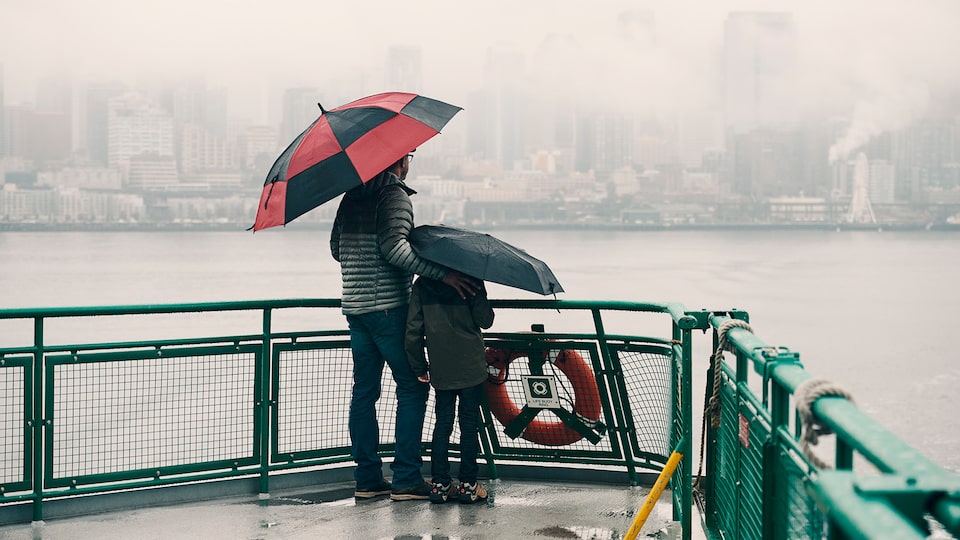 A father and son stand on the deck of a car ferry in the rain, holding umberalls and looking out at Seattle.