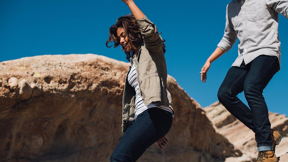 A young man and woman climb over large boulders with a bright blue sky behind them.