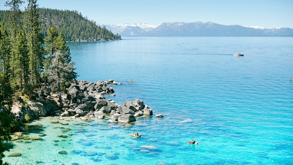 A very clear blue lake with rocks and trees along the shore and mountains in the distance.