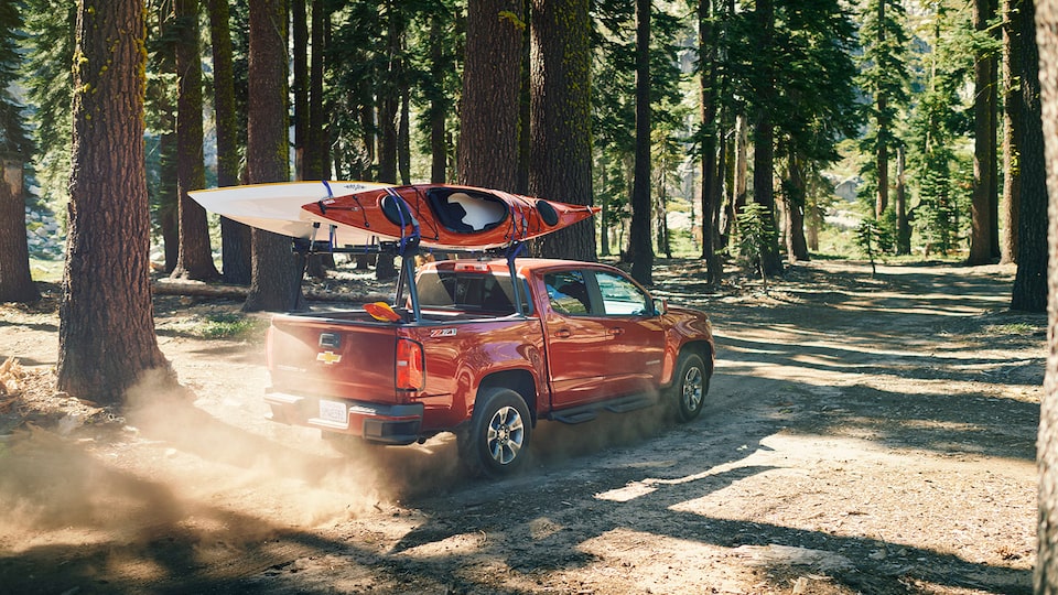 A Chevy Colorado with two kayaks on its rack drives through the forest.