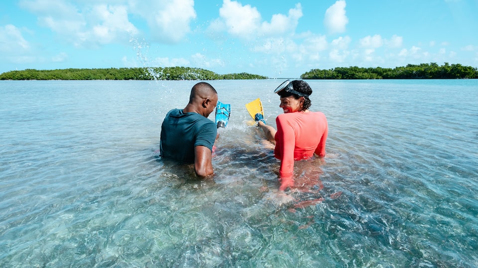A man and woman wearing swim flippers sit in shallow ocean water with islands in the background.
