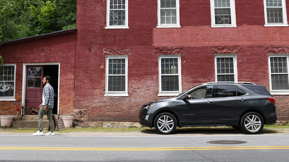 A Nightfall Gray Metallic Chevrolet Equinox sits on a street outside an old red brick two-story building. A man is walking down the street away from the Equinox.