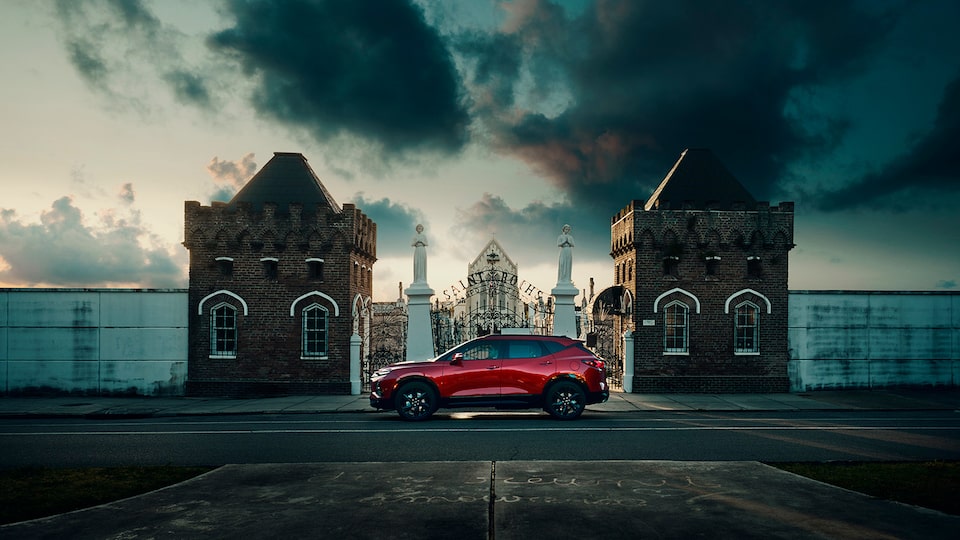 A red Chevy Blazer seen in profile on a street in front of a wrought iron gate between two large brick pillars with a cemetery in the background and dark clouds overhead.