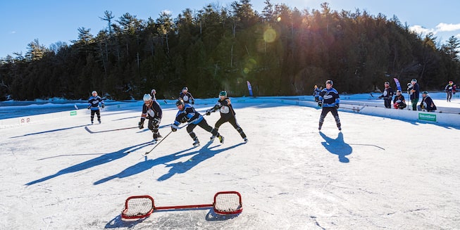 A group of people playing hockey on a frozen pond with a short goal net in the foreground and trees in the background.