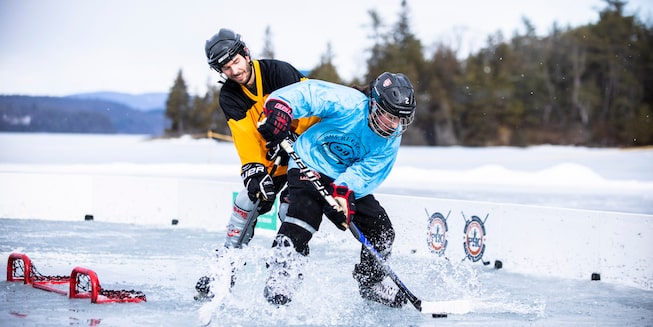 Two people in hockey gear compete for the puck on a frozen pond with trees in the background.