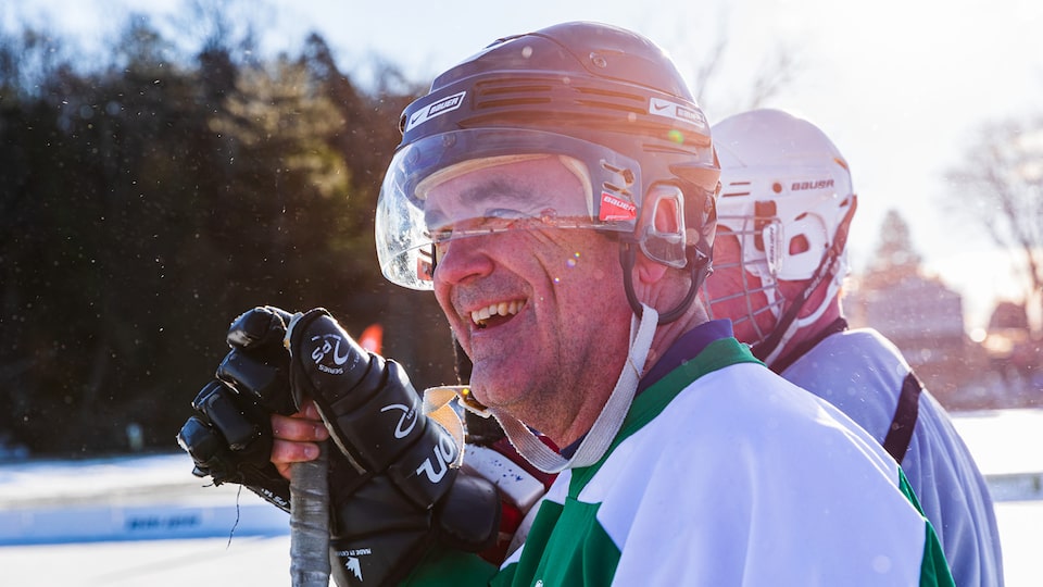 A man in a hockey helmet and gloves holding a hockey stick smiles.