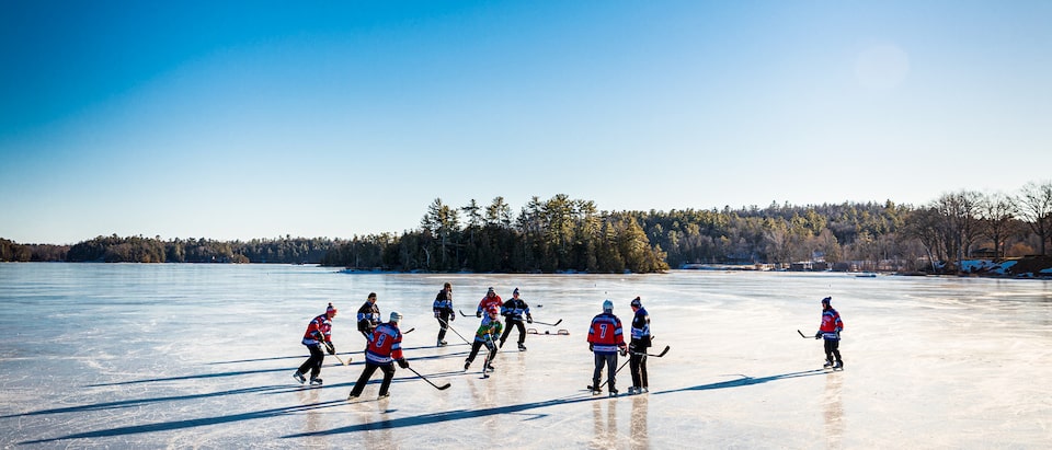 A group of people playing hockey on a frozen pond with a clear blue sky and trees in the background.